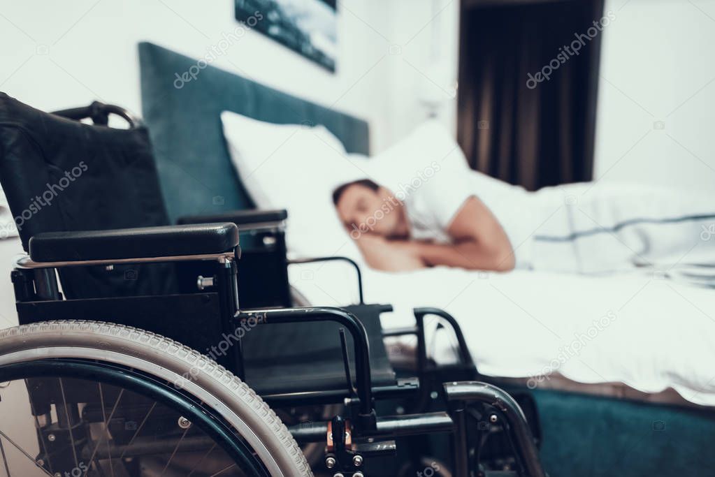 Disabled Person Sleeps Near Black Wheelchair. Closeup of Black Modern Invalid Chair on Foreground. Handicapped Young Person Lying in Large Bed With White Linens in Bright Bedroom