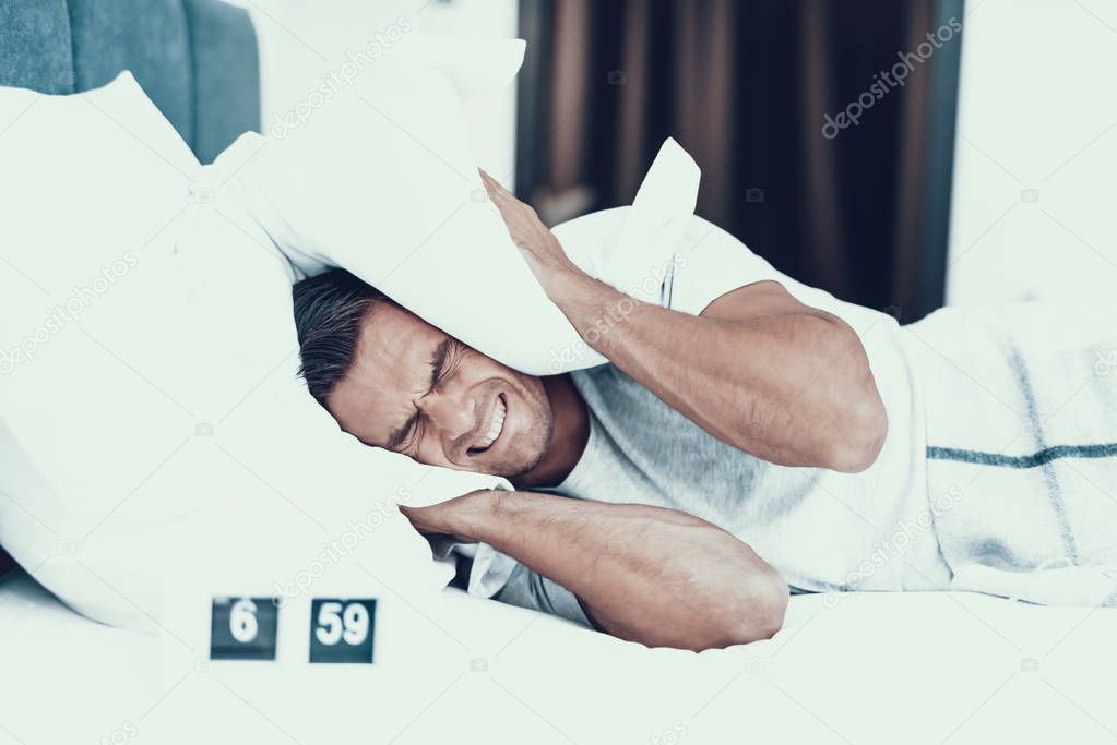 Angry Man Lies in White Bad and Hates Alarm Clock. Person in the Bedroom of Modern Apartment Wearing White T-shirt Hardly Waking Up and Closed Ears With Pillows. Morning Concept