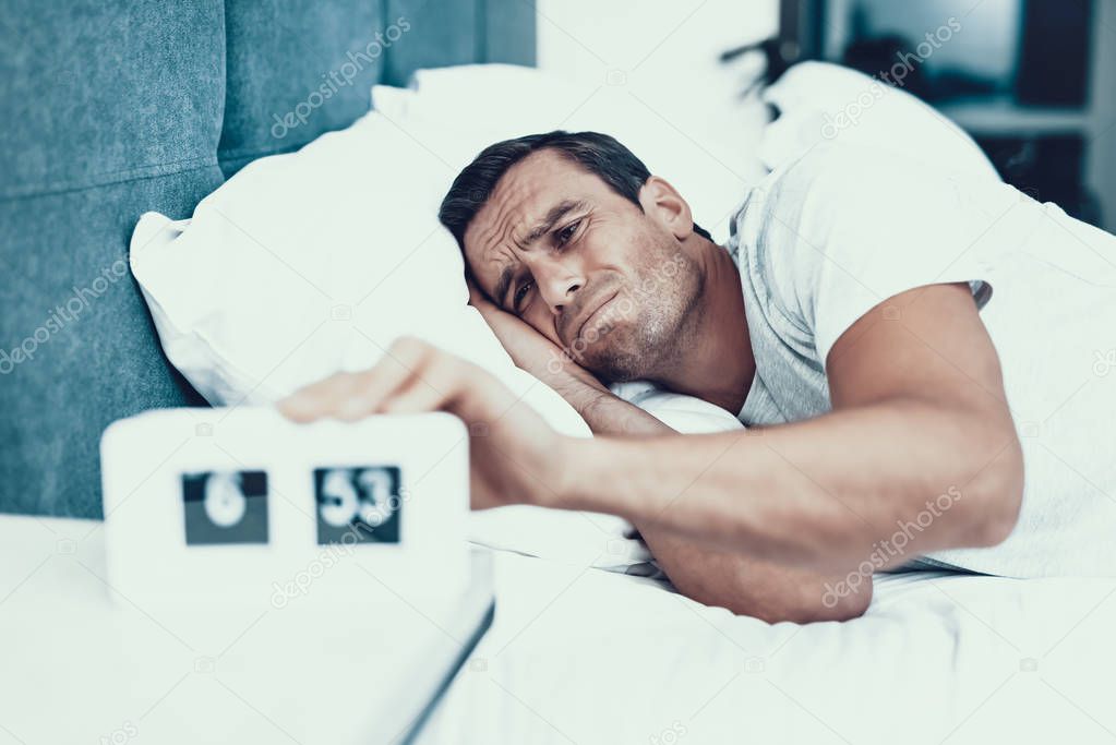 Man Hardly Awakes and Pulls Alarm Off in Morning. Handsome Young Person in Bad Mood Lying in Bedroom in Bed With White Linens Near Alarm Clock and Wearing White T-shirt. Morning Concept