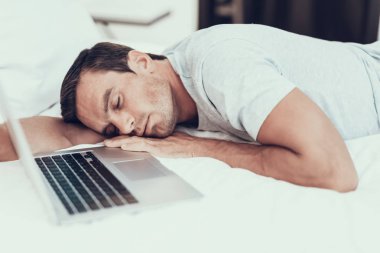 Adam Asleep dizüstü bilgisayarda çalışırken yatağa düştü. Beyaz çarşaflar hafif renkli tişört giyen ile büyük yatakta açılan not defteri yakınındaki yatan yorgun yakışıklı kahverengi saçlı genç insan portresi