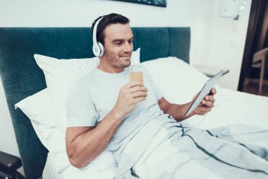 Adam Tablet ve beklemeler çay bardağı müzik dinler. Yakışıklı neşeli gülümseyen beyaz kişi parlak yatak beyaz keten ile yatakta rahatlatıcı. Adam kulaklıklar ayrı tutma Tablet ve içecekler kahve