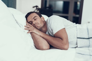 Alarm beyaz keten ile yatakta yakınındaki kişi uyur. Eller başının altına tutmak ve beyaz T-shirt giyen artalanları üzerinde bulanık tekerlekli sandalye ile yatak odasında yatan yakışıklı genç adam. Sabah kavramı