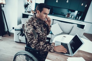 Tekerlekli sandalye orduda Laptop ve telefonlar üzerinde çalışmak. Üniformalı ciddi özürlü adam masada oturur ve Modern geçersiz sandalyede oturma odasında Park ve telefonu konuşurken oturan defter kullanır