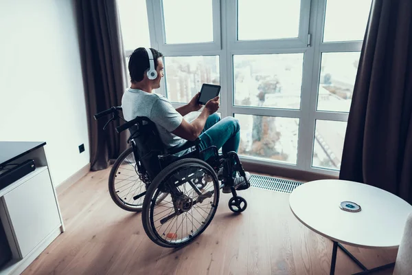 Smiling Disabled Man in Headphones Uses Tablet. Portrait of Handsome Happy Person in Casual Clothes Sits in Modern Wheelchair Parked near Window in Living Room or Hospital and Work on Touchscreen Pad