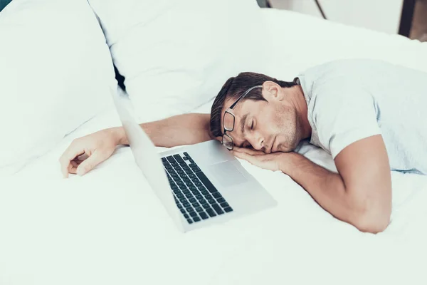 Man in Glasses Asleeps while Working on Laptop in Bed. Portrait of Exhausted Handsome Brown Haired Young Person Lying Near Opened Notebook in Big Bed with White Linens Wearing Light Coloured T-shirt