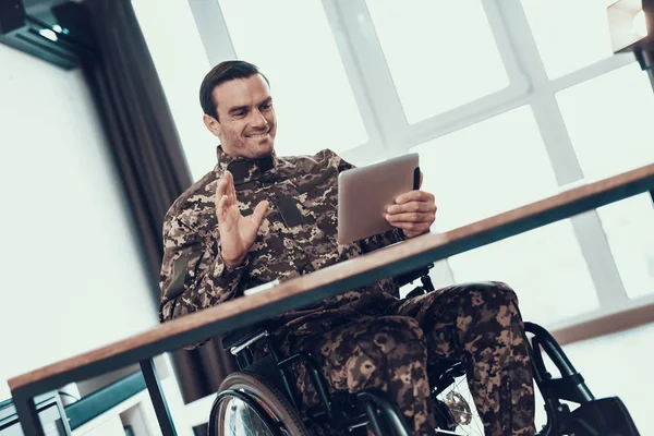 Military Man in Wheelchair Communicates via Video. Smiling Handsome Disabled in Military Uniform Works on Tablet at Desk Sitting in Modern Invalid Chair Parked in Bright Room and Shows Hello Gesture