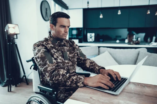 Military Man in Wheelchair Work on Laptop Indoors. Disabled in Military Uniform at Desk Uses Notebook. Man Sitting in Modern Invalid Chair Parked in Bright Living Room Apartment