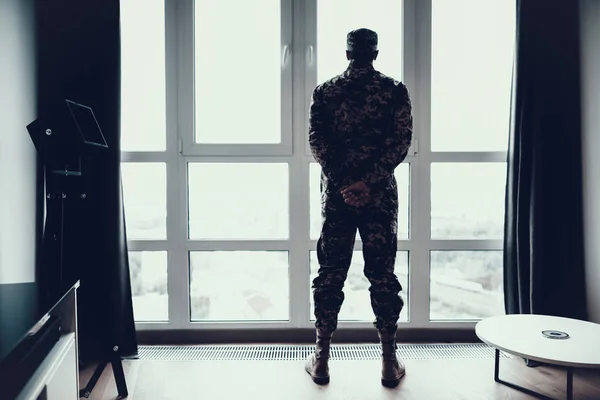 Back View of Man in Military Uniform Near Window. Serious Thoughtful Person Standing in Modern Apartment with Arms Crossed Behind Back and Looking at Large Panoramic Window Soldiery Concept