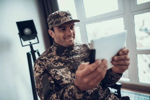 Disabled Military Man Uses Tablet in Wheelchair. Portrait of Caucasian Man Wearing Military Uniform Sitting in Modern Invalid Chair Parked near Large Panoramic Window in Living Room Apartment