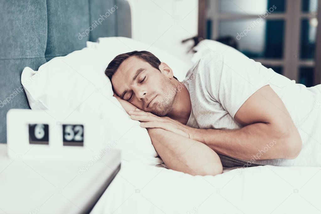 Person Sleeps Near Alarm in Bed With White Linens. Handsome Young Man Lying in Bedroom in Modern Apartment Keeping Hands Under Head and Wearing White T-shirt. Morning Concept