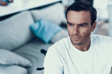 Özürlü düşünceli bakışla closeup. Yakışıklı ciddi beyaz kahverengi saçlı adam portresi kapalı sakin yüz ifade gösterilen beyaz T-Shirt giyen kanepe yakınındaki tekerlekli sandalyede oturur.