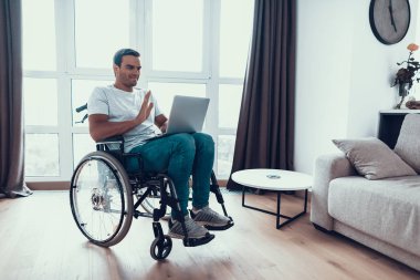 Kulaklığımda Engelli adam Laptop yolu ile iletişim kurun. Yakışıklı gülümseyen kişi Video yolu ile konuşmak ve Merhaba jest gösterilen ışık Modern salonunda tekerlekli sandalyede rahat kıyafetler oturur giyer.