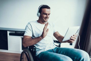 Kulaklığımda Engelli adam Tablet iletişim. Yakışıklı gülümseyen kişi T-Shirt giyiyor ve tekerlekli sandalye Internet tarama ve Video yolu ile konuşmak ve Merhaba jest gösterilen kot oturur.