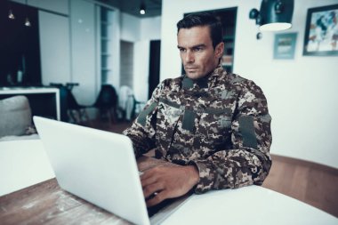 Yakışıklı askeri adam masada dizüstü bilgisayarda çalışıyor. Beyaz ciddi özürlü masasında askeri üniformalı gri defter kullanır. Modern aydınlık oturma odası dairede oturan adam