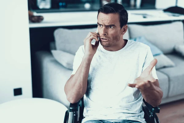 Disabled Person Has Serious Phone Conversation. Closeup Portrait of Caucasian Brown-haired Man Sits Indoors in Wheelchair Communicates Via Smartphone Wearing White T-Shirt Showing Sad Face Expression