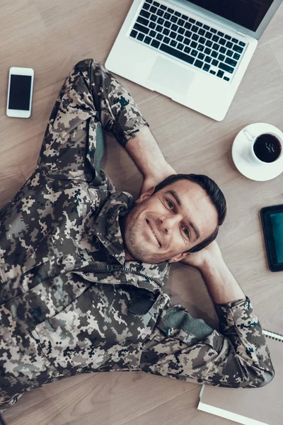 Military Man Lying on Table With Modern Gadgets. Cheerful Smiling Handsome Brown Haired Person in Military Uniform Surrounded by Laptop, Tablet, Smartphone and Cup of Coffee