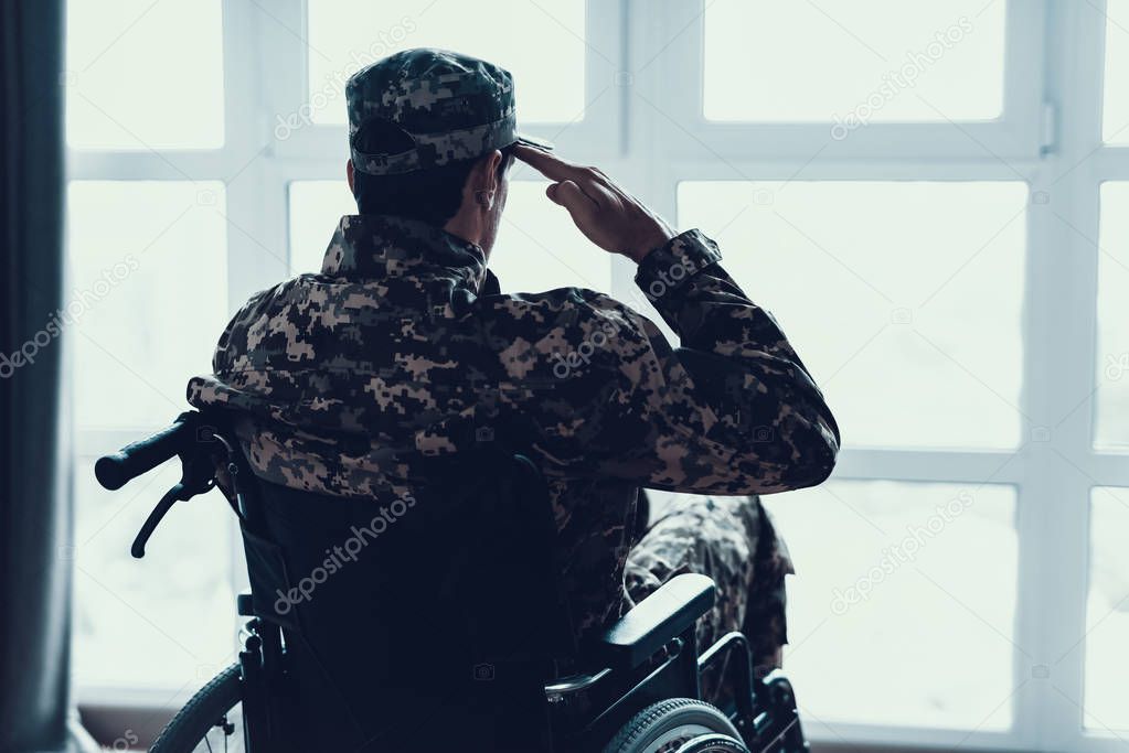 Disabled in Military Uniform Salutes in Wheelchair. Back View of Caucasian Man Wearing Hat Sitting in Modern Invalid Chair Parked near Large Panoramic Window in Living Room Apartment