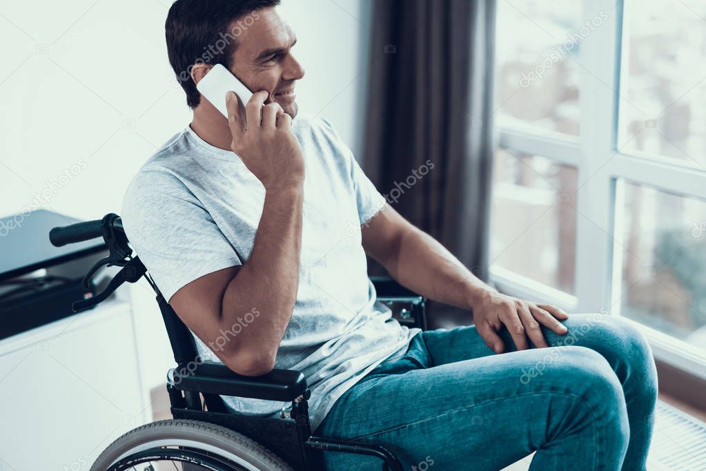 Handicapped Person in Wheelchair Talking Phone. Happy Caucasian Brown-haired Man Sits Indoors Near Window Wearing White T-Shirt. Male Communicates Using Smartphone and Keeps Hand on Knee