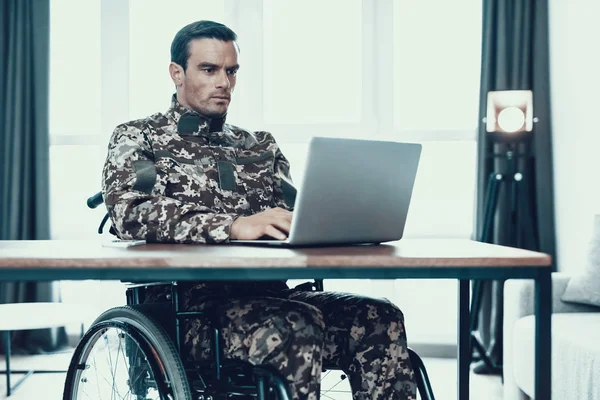 Military Man in Wheelchair Works on Laptop. Serious Disabled in Military Uniform at Desk Uses Gray Notebook. Man Sitting in Modern Invalid Chair Parked in Bright Living Room Apartment