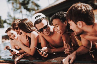Genç gülümseyen arkadaşlar havuz kenarında Smartphone kullanarak. Grup Genç insanlar mutlu konuşuyor ve havuz, Otel yüzme havuzu, cep telefonları kullanarak. Yaz tatili kavramı. Havuz partisi