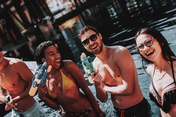 Young Smiling People in Pool with Water guns. Young Happy Friends standing Together with Colorful Water guns in Outdoor Swimming Pool. Friends at Pool Party. Summer Vacation Concept