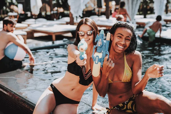 Two Young Smiling Women in Bikini at Poolside. Young Happy Friends holding Water Guns and Having Fun in Hotel Pool. Beautiful Women in Bikini Enjoying Outdoor Pool Party. Summer Vacation Concept