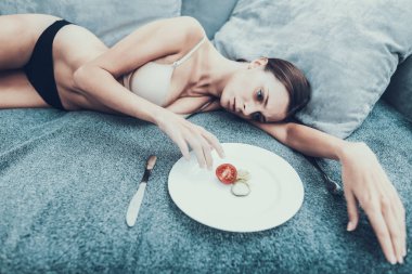 Slim Girl with Anorexia Lying on Sofa with Plate. Sliced Vegetables. Woman with Anorexia. Weigth Problem Concept. Tomato and Cucumber. Woman in Bra. Girl on Diet. Woman on Gray Sofa. clipart