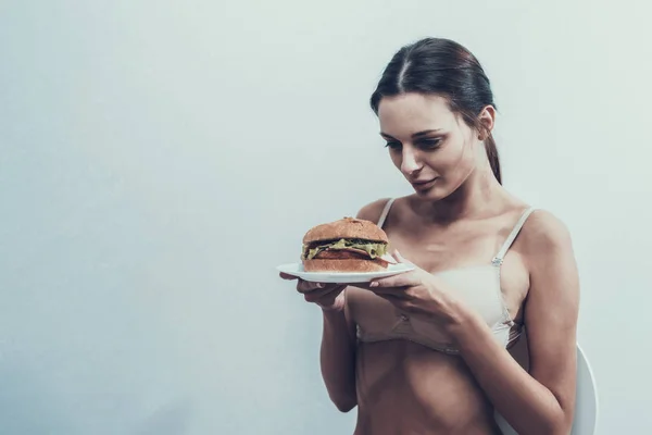 年轻的厌食症女孩在白色胸罩与汉堡 厌食症的女人 不健康的生活方式概念 不健康的食物 女孩与白板 饮食和保健概念 在白色房间的妇女 — 图库照片