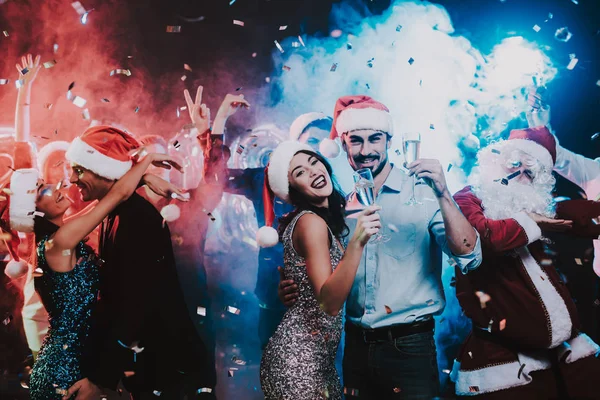 Happy Young People Dancing on New Year Party. Santa Claus. People in Red Caps. Happy New Year Concept. Glass of Champagne. Celebrating of New Year. Young Woman in Dress. Men in Suits.