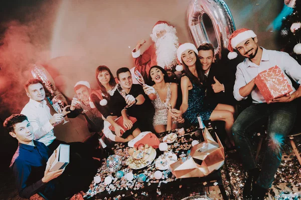 Young Happy People Celebrating New Year at Table. Santa Claus Costume. People in Red Caps. Happy New Year Concept. Bottle of Champagne. Celebrating of New Year. Man with Gift Box.