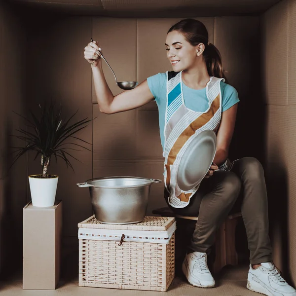 Young Woman in Apron Cooking in Cardboard Box. Life in Little Cardboard Box. Uncomfortable Life Concept. Personal Spase Concepts. Sitting Girl. Girl in Apron. Cooking Food. Smiling Woman.