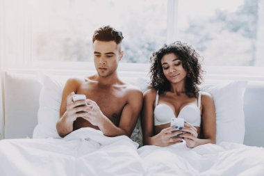 Genç güzel yatakta kullanarak Smartphones Çift. Çekici kadın ve yan yana yatakta yatarken ve cep telefonu kullanma yakışıklı adam. Mutlu sevimli çift iç çamaşırı evde yatakta rahatlatıcı.
