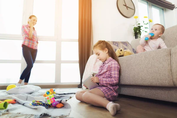 お母さん電話の話中の部屋で遊んでいる子供たち リビング ルームの大パノラマの窓近くに母が立っています 赤ちゃんグッズをおいて口と床にタブレットを使用してより古い子供のソファに座って — ストック写真