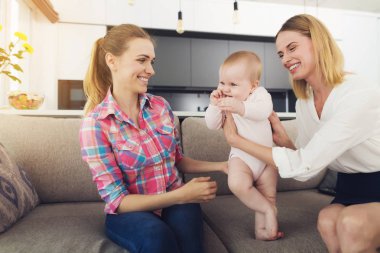 Koltukta oturan çocuk bakıcısı için veren çocuk anne. Gülümseyen güzel beyaz anne Holding şirin güzel bebek beyaz Bluse oturma odasında rahat ışık kanepede oturan giymiş elinde