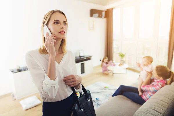 女商人妈妈在客厅里会说话的电话 保姆在背圆上看孩子 而妈妈则在电话里 美丽的高加索金发女郎母亲穿着白色的毯子拿着黑色的袋子 — 图库照片