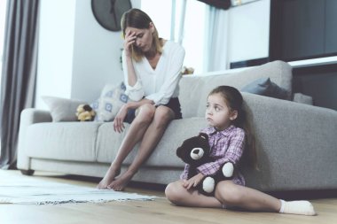 Girl Sits on Floor with Teddy Bear Near Upset Mom. Disappointed Caucasian Young Mother Sitting on Light Couch in Modern Living Room Holds Head. Bad Family Relationship Concept clipart