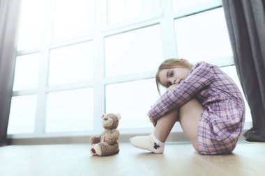 Oyuncak ayı katta kapalı ile üzgün kız Sit. Kameraya bakarak büyük panoramik pencere yakınındaki sıkışık diz ile oturan rahat kıyafetler giymiş çocuk oldukça üzgün. Çocukluk kavramı