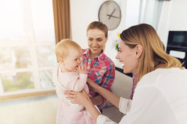 Babysitter and Mother Playing with Baby Indoors. Smiling Caucasian Happy Nanny Holding Adorable Baby and Cheerful Mom Taking Cute Daughter on Hands in Bright Living Room at Home clipart