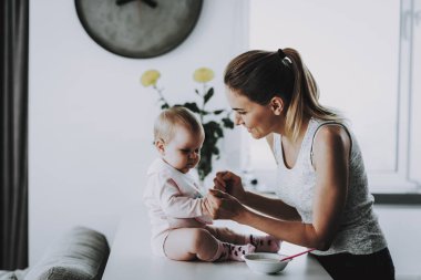 Evde mutlu gülümseyen anne beslemeleri çok güzel evlat. Bright salon şirin aç yürümeye başlayan çocuk sahip odaya kase annelik ve çocukluk kavramı yakınındaki masada otururken anne ile