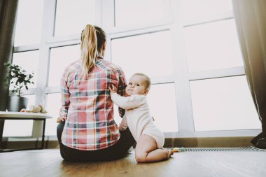 Annem oturma arka küçük çocuğun pencerenin yanında. Portre tatlı güzel beyaz çocuk duran, Modern oturma odası Genç Anne giyiyor rahat kıyafetler yakınındaki. Annelik ve çocukluk kavramı