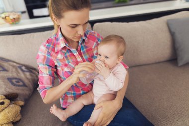 Bebek şişeden içer ve diz anneler üzerinde oturur. Mutlu anne rahat ışık kanepede oturan ve oturma odasında güzel Beyaz Toddler beslenme rahat kıyafetler giyer. Annelik kavramı