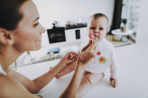 Happy Mom Feeds Adorable Baby with Spoon Indoors. Mother Diving Healthy Food to Cute Hungry Toddler In Bright Living Room. Happy Family Having Breakfast in Kitchen. Motherhood and Childhood Concept