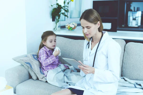 Young Female Doctor Hold Pad and Make Diagnosis. Pretty Little Ill Girl Having Cold Sitting on Couch with Nurse Wearing Stethoscope and White Medical Gown Looking at Tablet Screen.