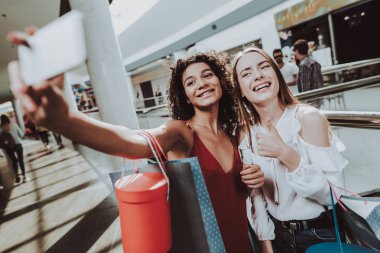 Selfie alışveriş merkezinde alarak paketleri iki kızla. Smartphone kullanarak. Dijital aygıt elinde. Alışveriş kavramı. Kırmızı elbiseli kız. Kadın beyaz gömlekli. Paketleri tutarak. Paketi elinde alışveriş merkezi.