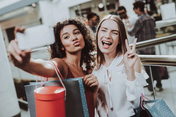 Two Girls with Packages Taking Selfie in Mall. Using Smartphone. Digital Device in Hand. Shopping Concept. Girl in Red Dress. Woman in White Shirt. Holding Packages. Pack from Mall in Hands.