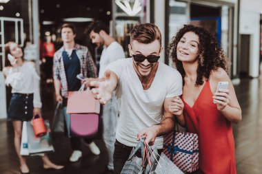 Genç paketleri ile alışveriş merkezinde alışveriş çift. Alışveriş kavramı. Kırmızı elbiseli kız. Tıraşsız genç adam tutma paketleri. Paketi elinde alışveriş merkezi. Gülümseyen insanlar. Erkek Güneş gözlüğü.