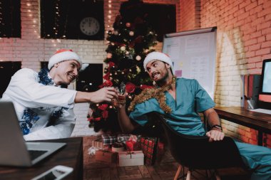 İki gülümseyen doktor ofisinde yılbaşı kutlamak. Santa Claus şapkalı adam. Ofiste Noel ağacı. Bilgisayar masasında. Beyaz ceketli adam. İçki içerken. Yeni yıl partisi. Meslektaşım ile kutlama.