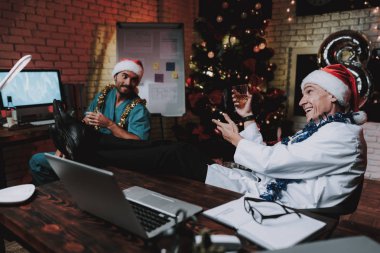 Colleageu fave eğlenceli ofis ile genç doktor. Yeni yıl arifesi. Ofiste Noel ağacı. Bilgisayar masasında. Beyaz ceketli adam. Yeni yıl kutlama. Dijital aygıt kullanarak. Santa Claus Cap.