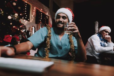 Colleageu fave eğlenceli ofis ile genç doktor. Yeni yıl arifesi. Ofiste Noel ağacı. Bilgisayar masasında. Beyaz ceketli adam. Yeni yıl kutlama. Dijital aygıt kullanarak. Santa Claus Cap.