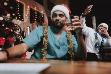 Colleageu fave eğlenceli ofis ile genç doktor. Yeni yıl arifesi. Ofiste Noel ağacı. Bilgisayar masasında. Beyaz ceketli adam. Yeni yıl kutlama. Dijital aygıt kullanarak. Santa Claus Cap.